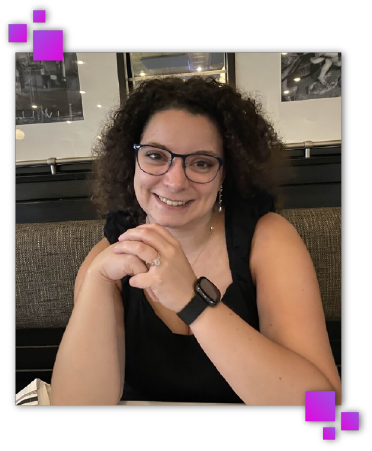 Website developer and professional branding expert, Dakota. In the photo she is smiling and holding her hands with her elbows on a table. She is a white woman with curly brown hair wearing a black watch, drop pearl earrings, and black glasses.